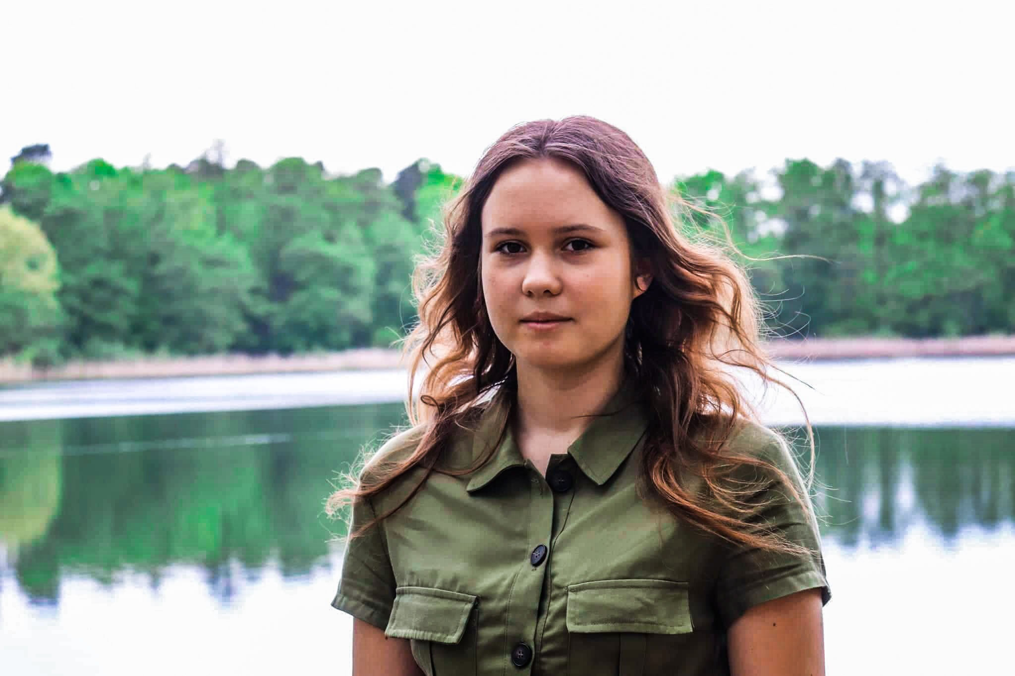 Portret młodej kobiety o brązowych włosach w zielonej koszuli na tle jeziora i lasu.