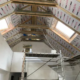 Poddasze w trakcie ocieplania, widoczne drewniane krokwie, izolacja termiczna JKO enertherm, okna dachowe i rusztowanie malarskie.