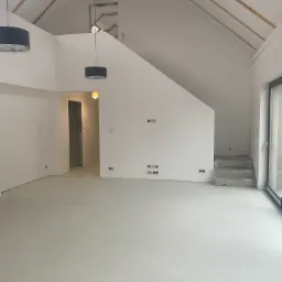 Surowe schody betonowe prowadzące na antresolę w przestronnym, jasnym pomieszczeniu z białymi ścianami i świeżo wylaną, jasną posadzką, widoczne drewniane elementy więźby dachowej i okno dachowe.