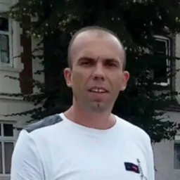 Portret mężczyzny w białej koszulce na tle budynku z oknami i zielenią, możliwe zdjęcie pracownika firmy remontowej lub budowlanej.