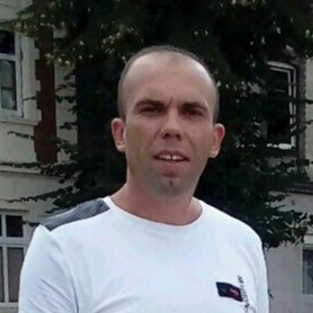 Portret mężczyzny w białej koszulce na tle budynku z oknami i zielenią, możliwe zdjęcie pracownika firmy remontowej lub budowlanej.