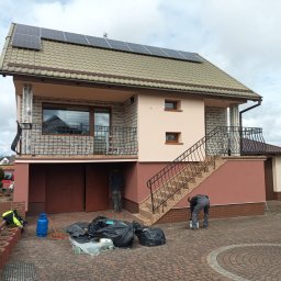 ŚWIECA INVEST ŁUKASZ ŚWIECZKOWSKI - Dwukondygnacyjny dom z panelami słonecznymi na dachu, w trakcie malowania elewacji zewnętrznej, widoczne worki na śmieci, butla gazowa i pracownicy przy pracy.