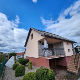 ŚWIECA INVEST ŁUKASZ ŚWIECZKOWSKI - Dwukondygnacyjny dom z beżową elewacją i balkonem z kutą balustradą, widok z ogrodu z ozdobnymi krzewami w słoneczny dzień.