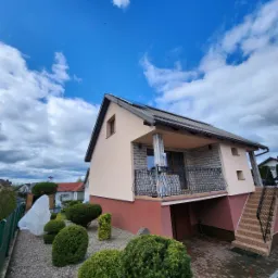 Dwukondygnacyjny dom z beżową elewacją i balkonem z kutą balustradą, widok z ogrodu z ozdobnymi krzewami w słoneczny dzień.