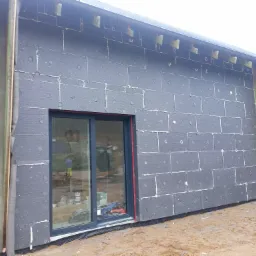 Budynek w trakcie ocieplania, widoczna czarna izolacja termiczna z szarymi kołkami, okno przesuwne z ciemną ramą, fragment elewacji z deskami i folią wiatroizolacyjną.