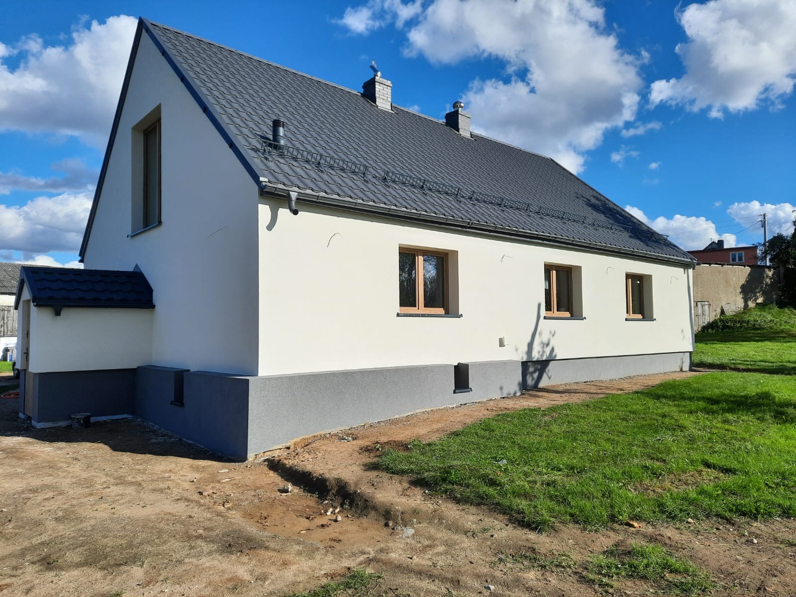 Nowo wybudowany dom jednorodzinny z szarą dachówką, jasną elewacją i szarym cokołem, widok z zewnątrz w słoneczny dzień.