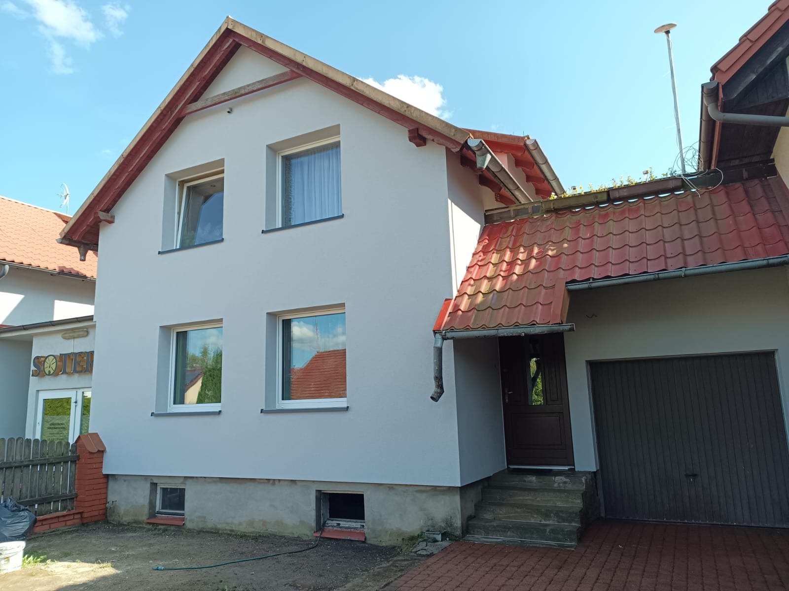 Dwukondygnacyjny dom z szarą elewacją, czerwonym dachem i widocznymi oknami, po remoncie, w słoneczny dzień.