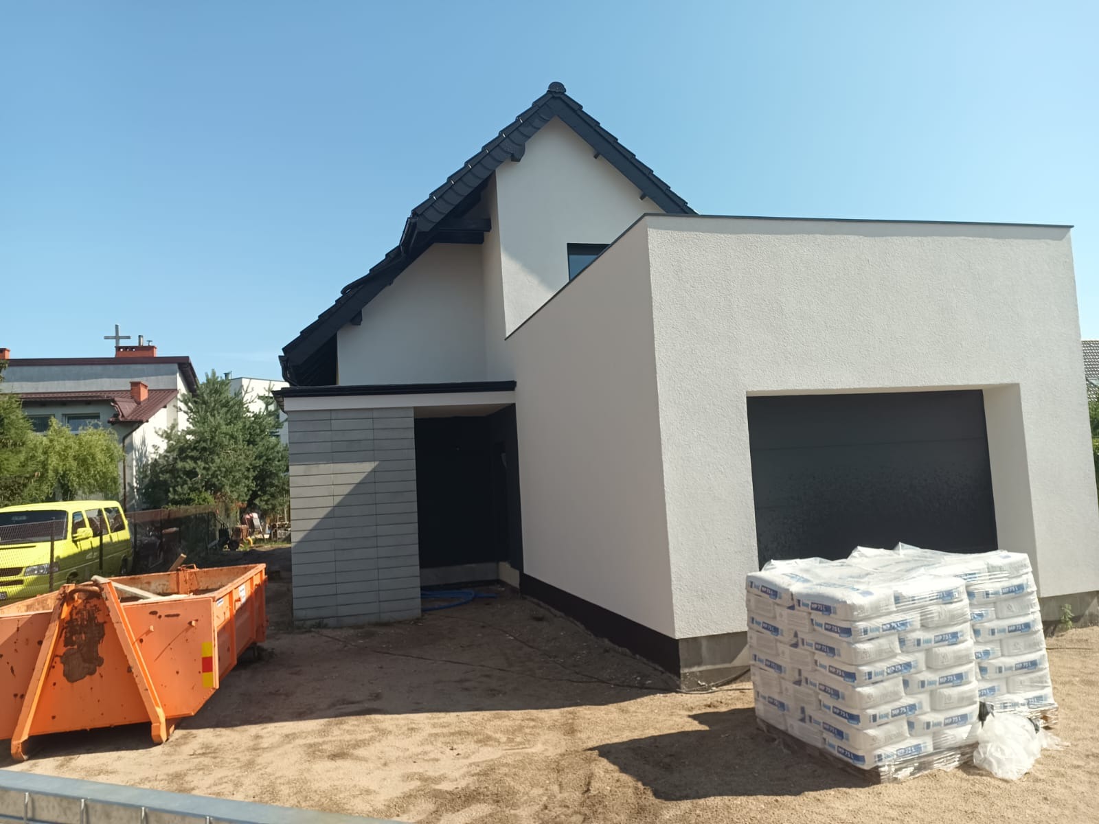 Nowoczesny dom jednorodzinny z białymi ścianami i czarnym dachem w trakcie budowy, widoczny pomarańczowy kontener na gruz i palety z workami zaprawy.