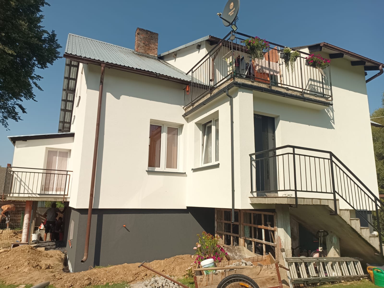 Dwupiętrowy dom z balkonem w trakcie malowania elewacji, z widocznym fragmentem rusztowania i pracownikami w tle.