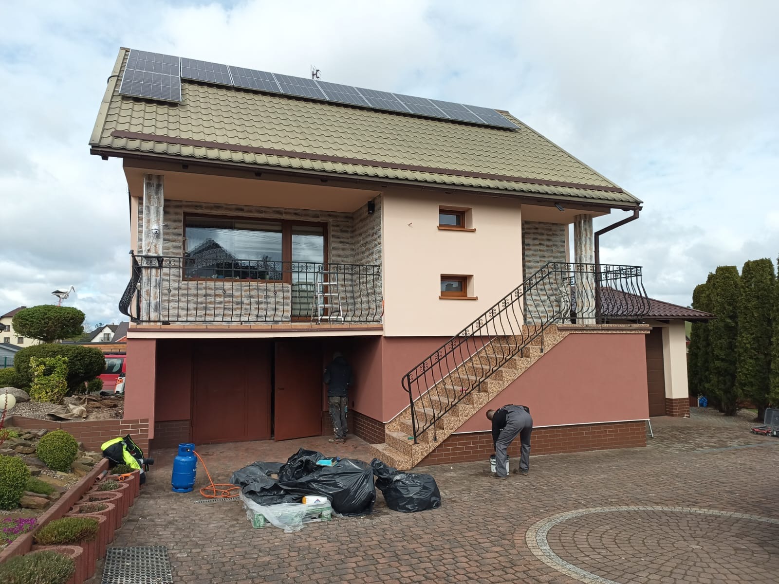 Dwukondygnacyjny dom z panelami słonecznymi na dachu, w trakcie malowania elewacji zewnętrznej, widoczne worki na śmieci, butla gazowa i pracownicy przy pracy.