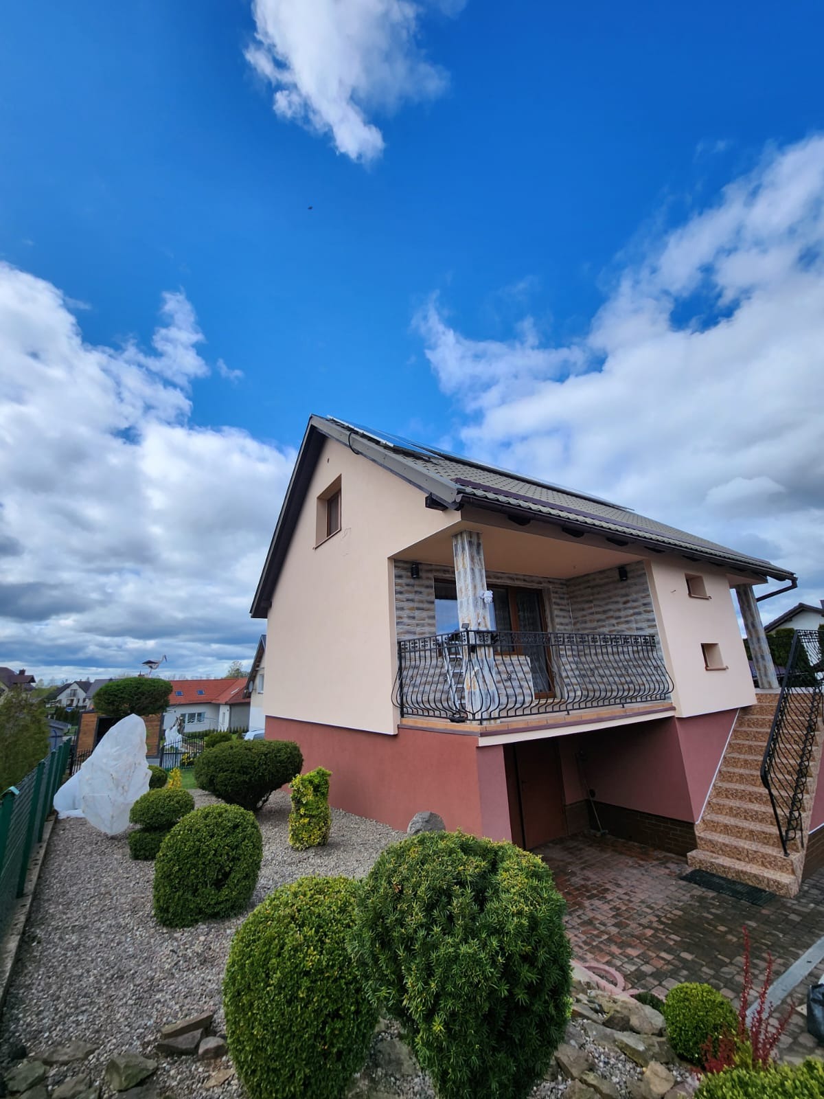 Dwukondygnacyjny dom z beżową elewacją i balkonem z kutą balustradą, widok z ogrodu z ozdobnymi krzewami w słoneczny dzień.