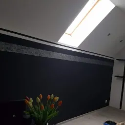 Ciemna ściana w pokoju na poddaszu z oknem dachowym, ozdobiona poziomym pasem z efektem dekoracyjnym, wazon z tulipanami w rogu.