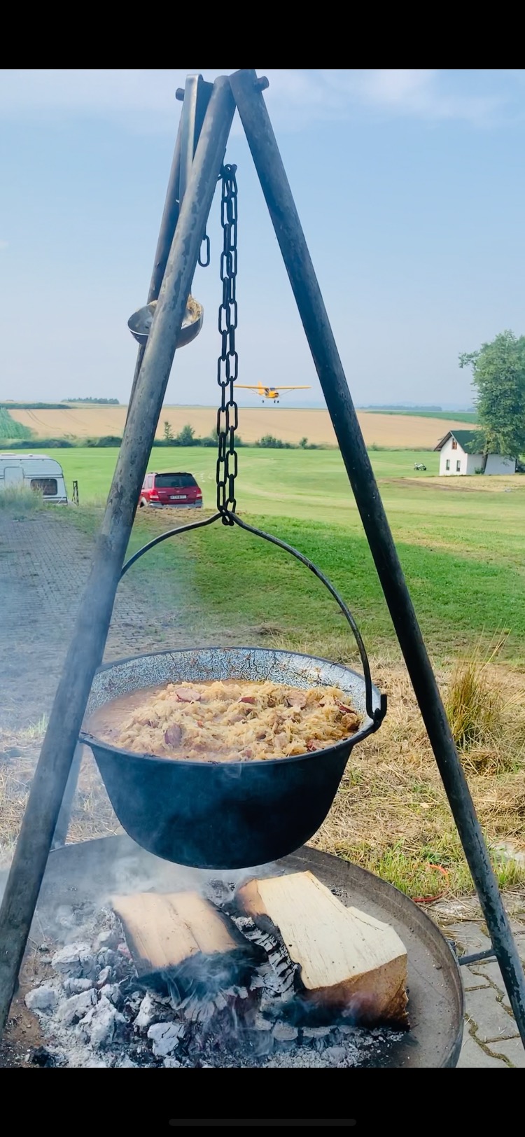 Duży kociołek z kapustą i kiełbasą gotowany na ognisku w plenerze, w tle samolot na pasie startowym.