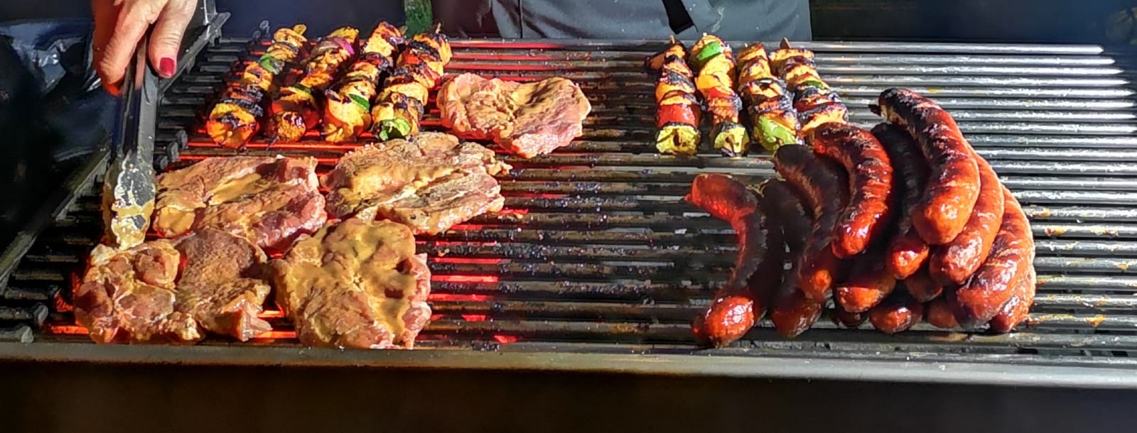 Grill z szaszłykami z kurczaka i warzyw, marynowanymi kawałkami mięsa oraz brązowiejącymi kiełbasami, przyrządzanymi nad żarzącym się węglem, obsługiwany szczypcami przez osobę z pomalowanymi...