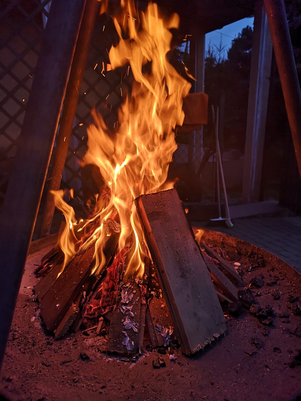Ognisko w metalowej misie, płomienie oświetlają drewniane deski i iskry, w tle drewniana konstrukcja i nocne niebo.