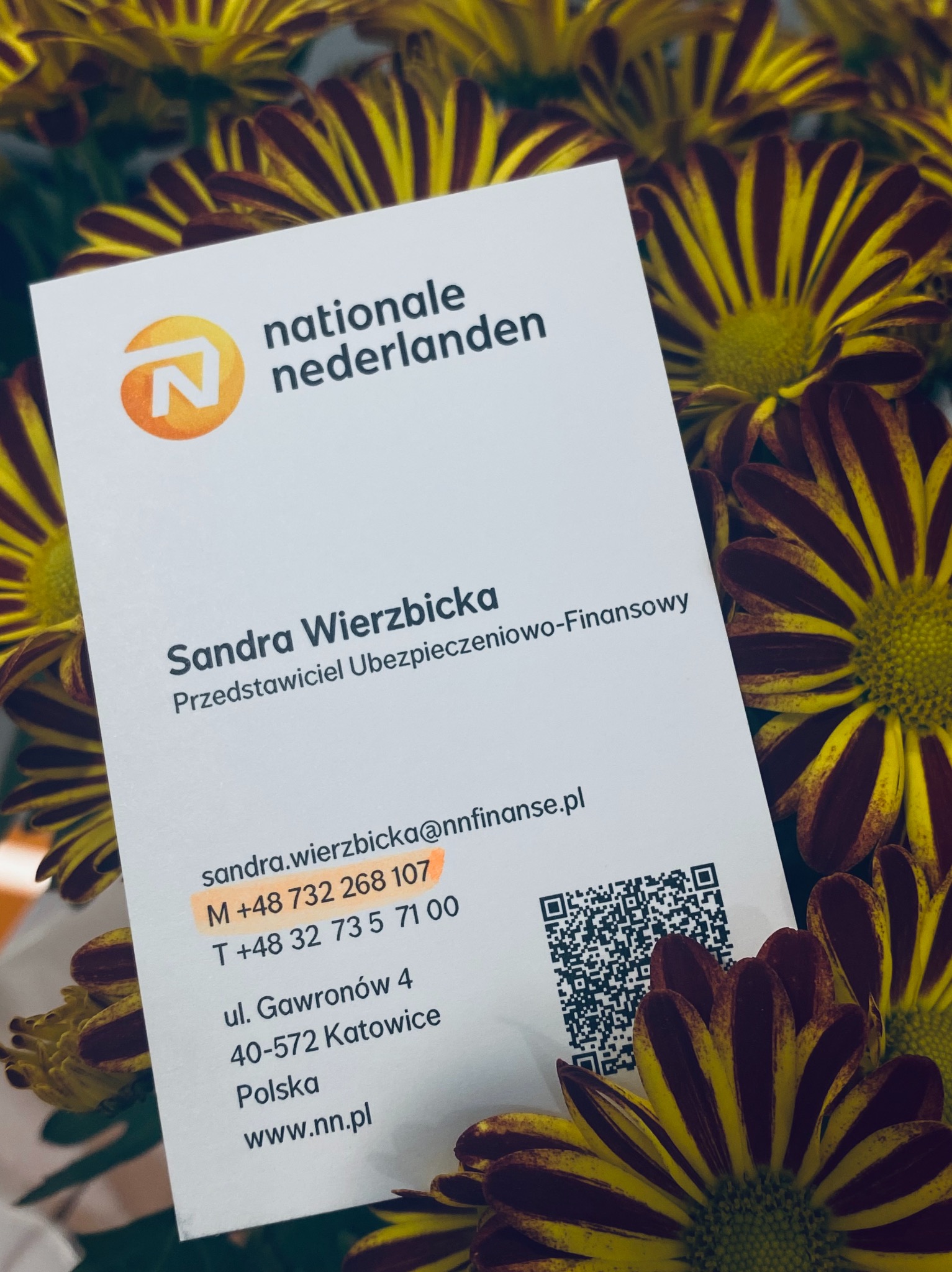 Wizytówka przedstawiciela firmy Nationale Nederlanden, Sandra Wierzbicka, na tle kwiatów. Wizytówka zawiera logo firmy, dane kontaktowe i kod QR.