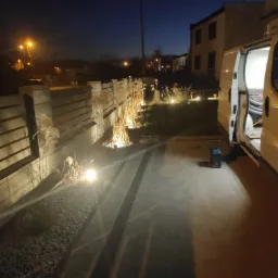 Oświetlony ogród wzdłuż betonowego ogrodzenia z ozdobnymi trawami i lampami punktowymi, widoczny samochód dostawczy z otwartymi drzwiami w wieczornej scenerii.