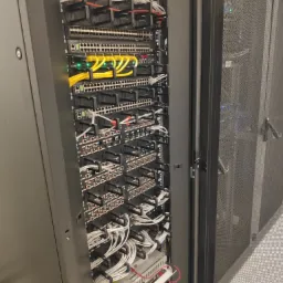 Szafa serwerowa z widocznymi przełącznikami sieciowymi, kablami Ethernet w kolorach żółtym i białym, uporządkowanymi za pomocą czarnych uchwytów; widoczna siatka drzwi szafy.