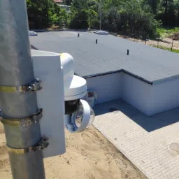 Zewnętrzna kamera monitoringu zamocowana na metalowym słupie za pomocą metalowych obejm, widok z góry na budynek z płaskim dachem i kostką brukową.