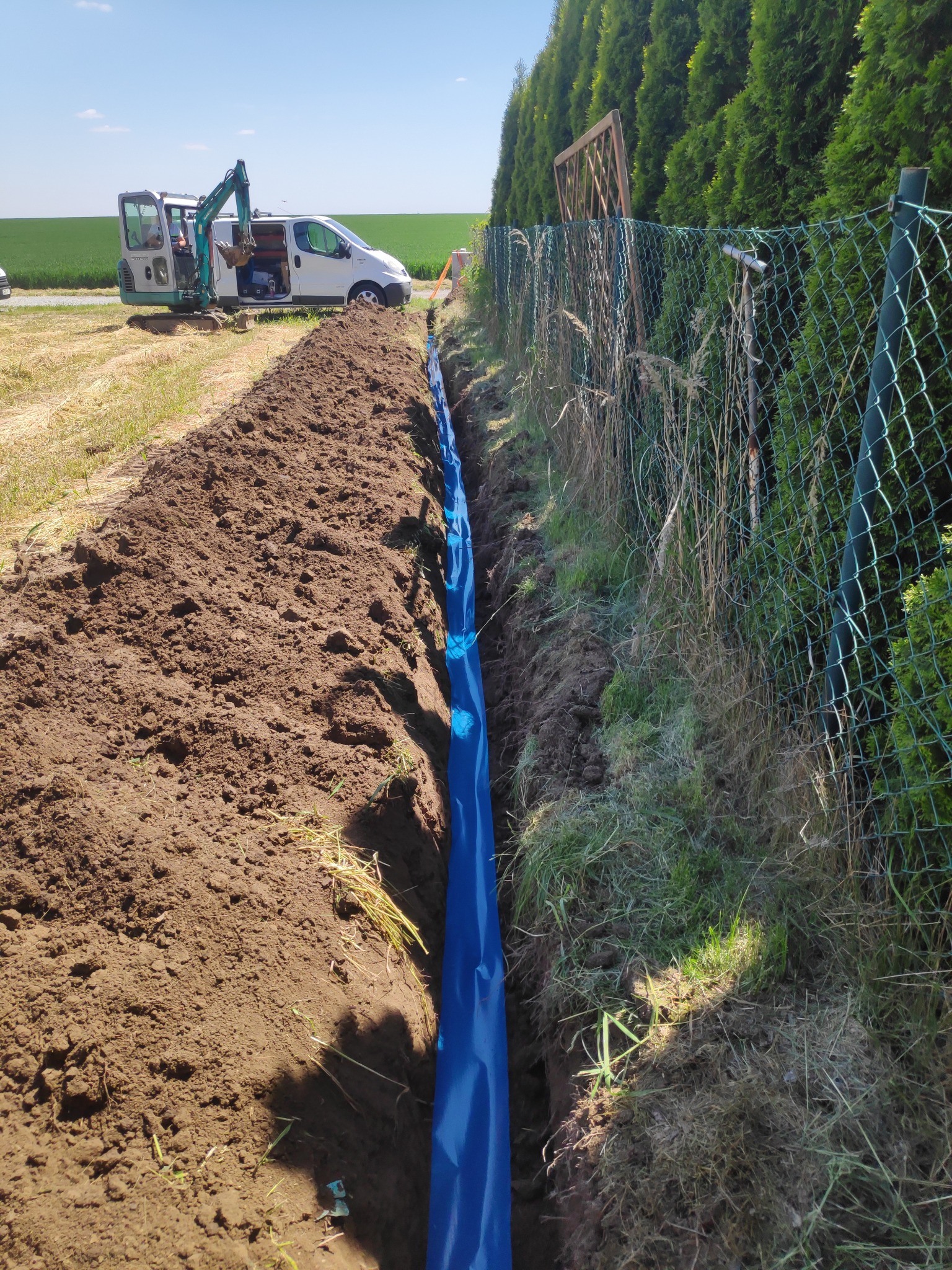 Wykopany rów z niebieską folią wzdłuż ogrodzenia z siatki, w tle minikoparka i biały samochód dostawczy na tle zielonego pola.