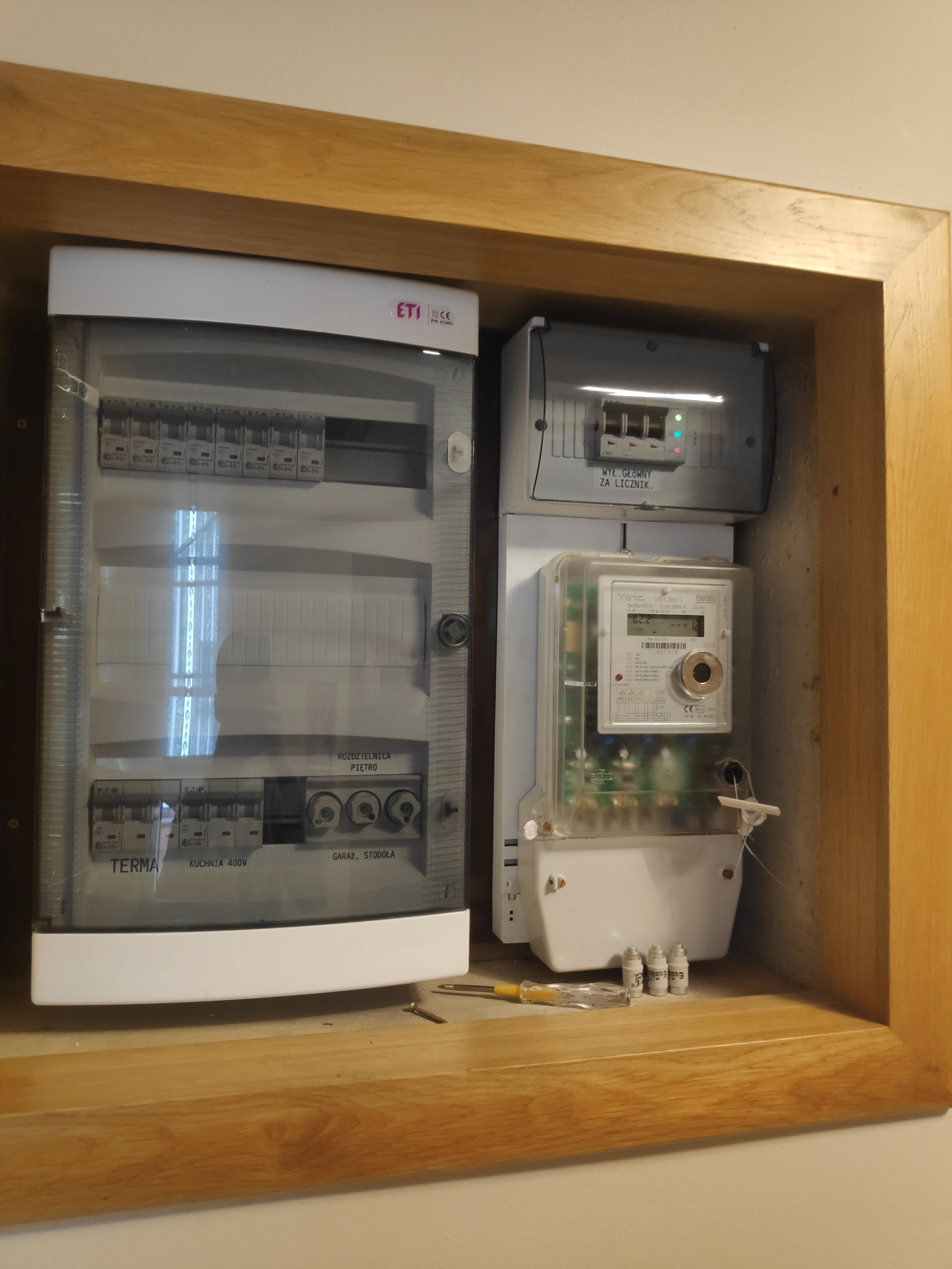 Rozdzielnia elektryczna z licznikiem i bezpiecznikami w drewnianej obudowie, widoczny śrubokręt i zapasowe bezpieczniki