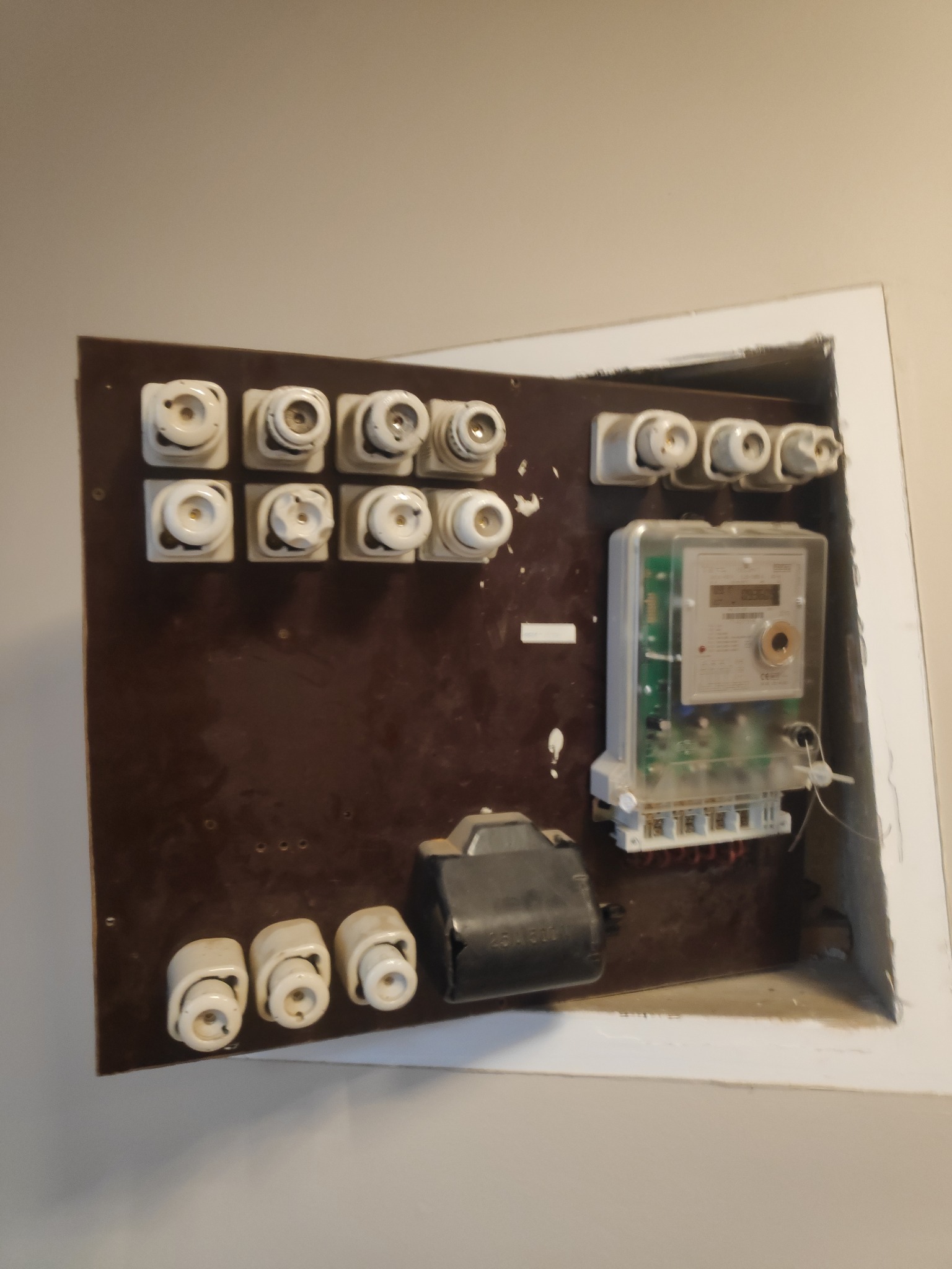 Otwarta skrzynka bezpiecznikowa z ceramicznymi bezpiecznikami topikowymi i licznikiem energii elektrycznej, widoczne okablowanie, instalacja wnękowa w ścianie.