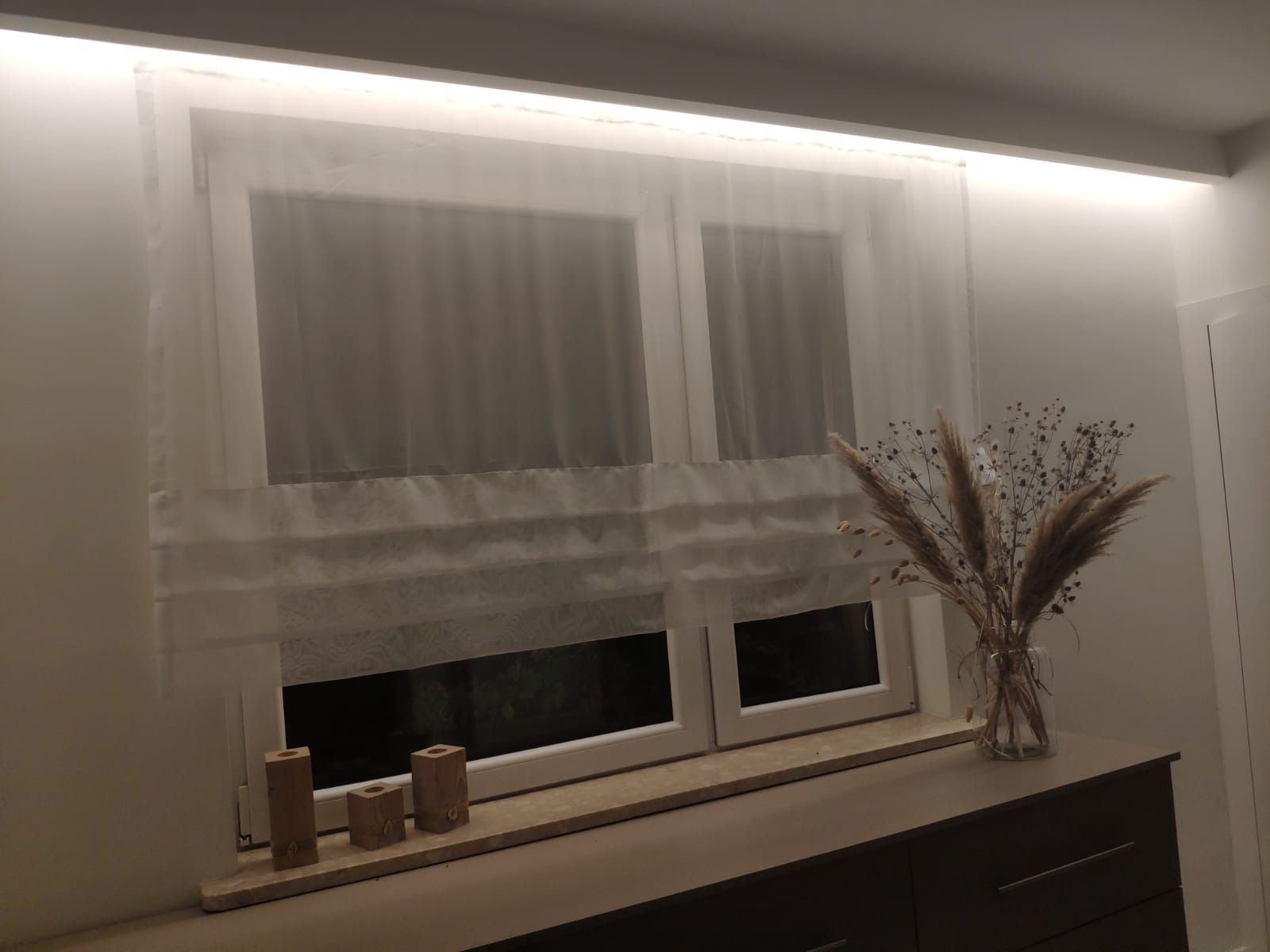 Okno z białymi, zwiewnymi roletami rzymskimi, podświetlone listwą LED, dekorowane drewnianymi świecznikami i bukietem suszonych traw w szklanym wazonie.