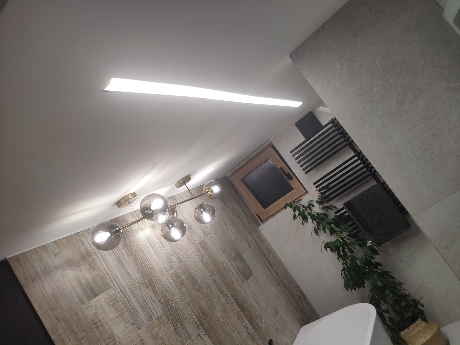 Nowoczesna łazienka z drewnopodobnymi płytkami ściennymi, szklaną lampą sufitową z kulami, grzejnikiem drabinkowym i rośliną w koszu.