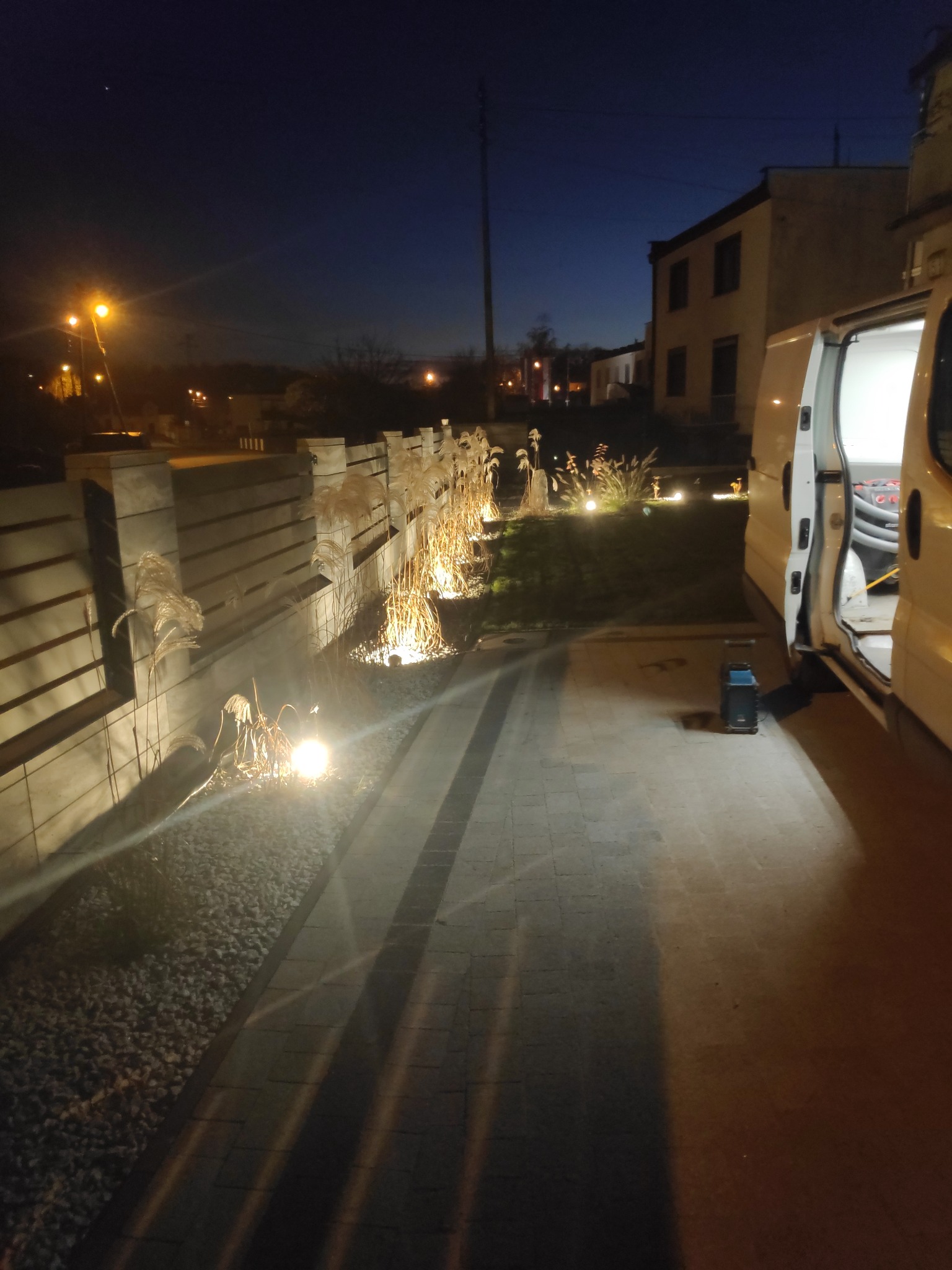 Oświetlony ogród wzdłuż betonowego ogrodzenia z ozdobnymi trawami i lampami punktowymi, widoczny samochód dostawczy z otwartymi drzwiami w wieczornej scenerii.