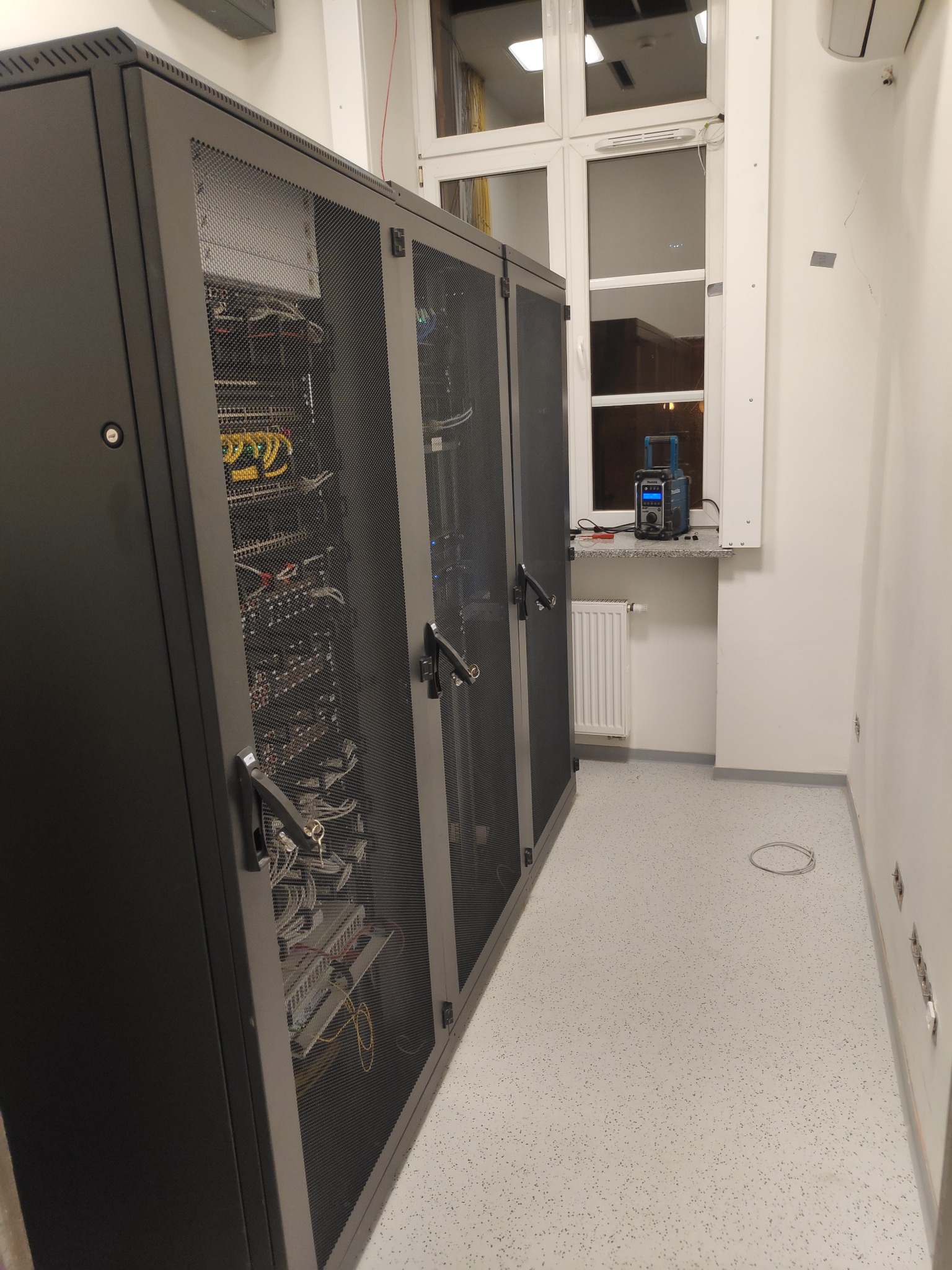 Trzy szafy serwerowe z widocznym okablowaniem i sprzętem wewnątrz, ustawione w jasnym pomieszczeniu z oknem, na parapecie narzędzie diagnostyczne.