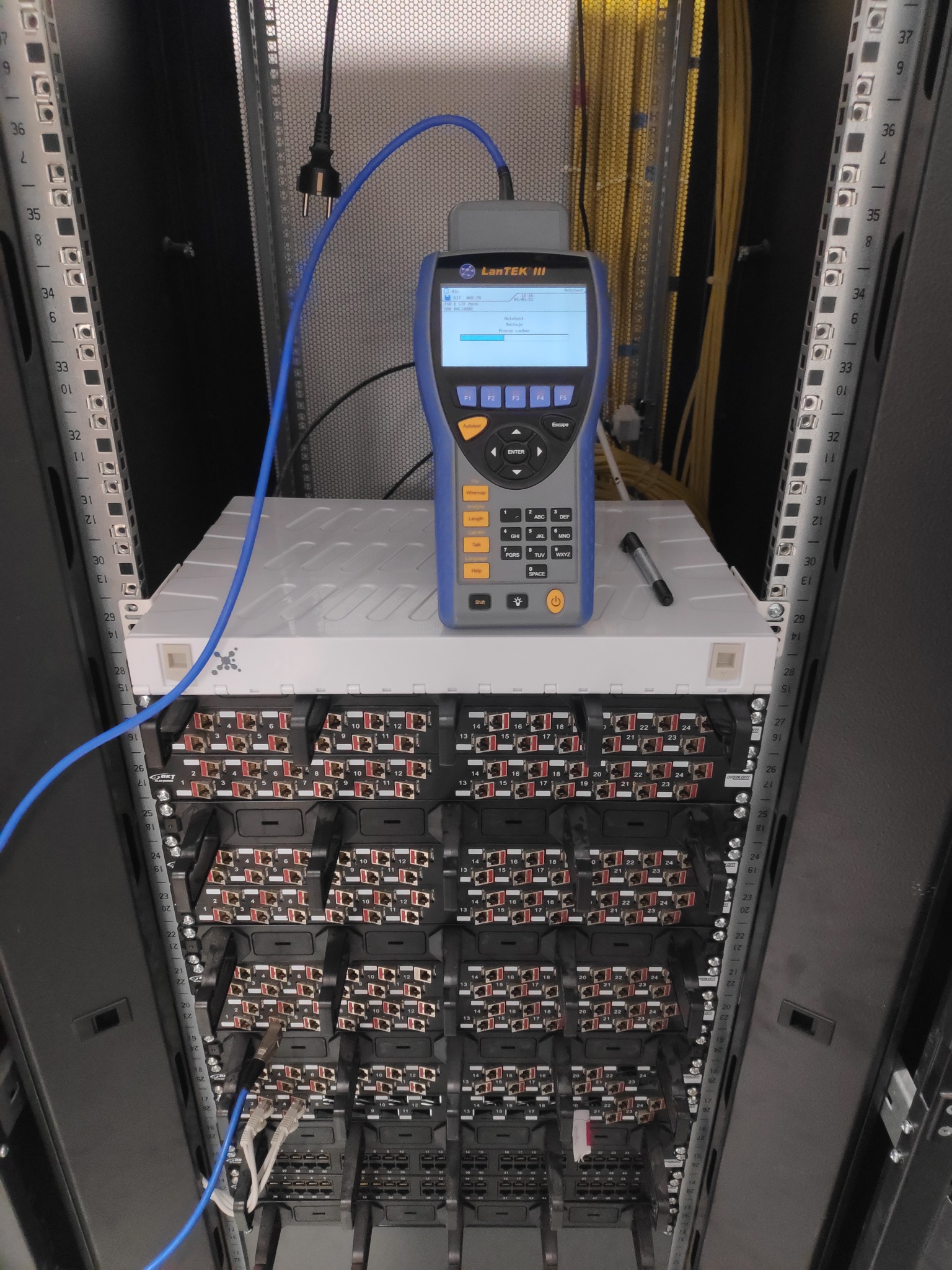 Tester LanTEK III podłączony niebieskim kablem do panelu krosowniczego w szafie rack, z widocznym markerem obok urządzenia, pomiar w toku.