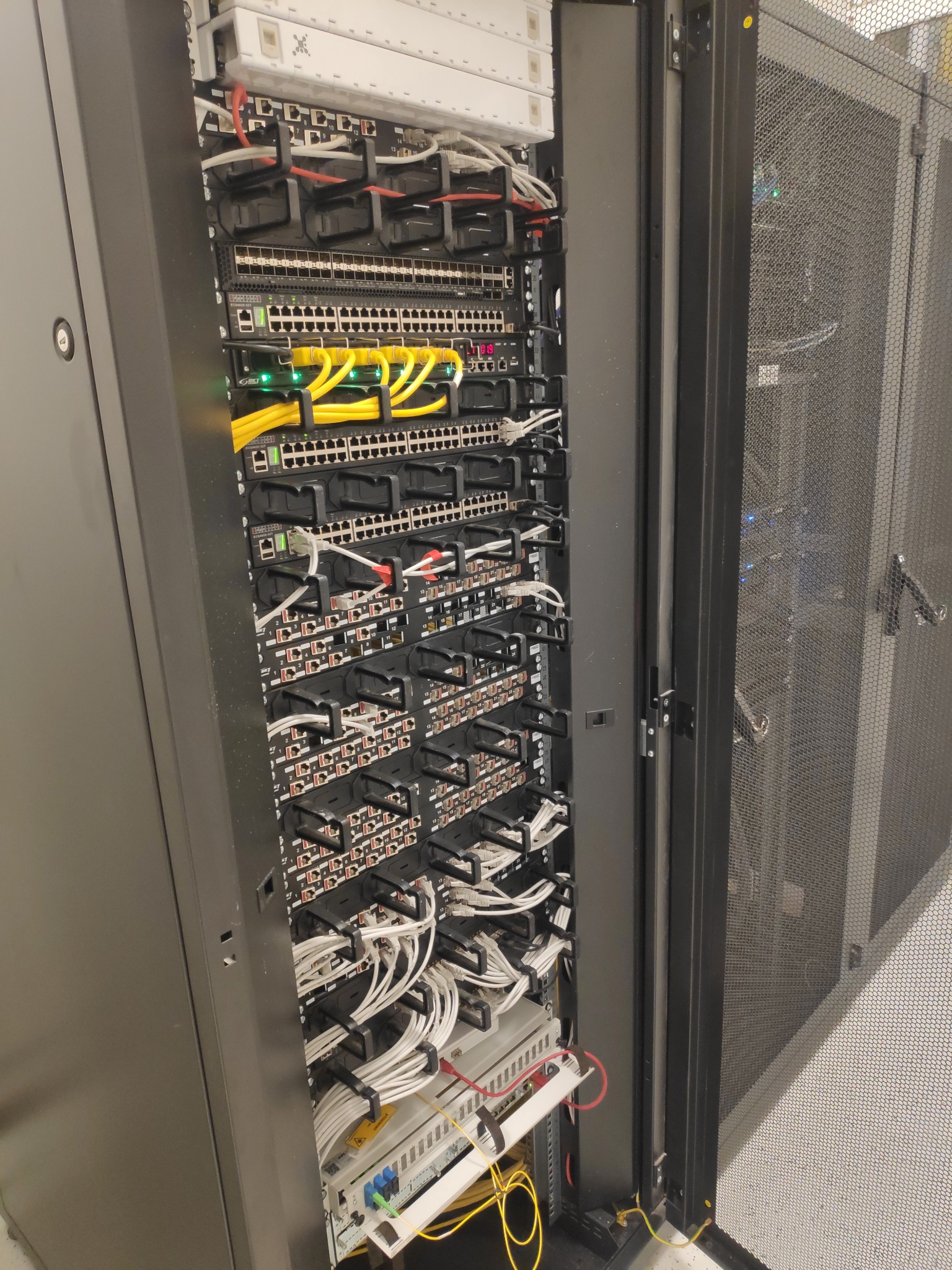 Szafa serwerowa z widocznymi przełącznikami sieciowymi, kablami Ethernet w kolorach żółtym i białym, uporządkowanymi za pomocą czarnych uchwytów; widoczna siatka drzwi szafy.