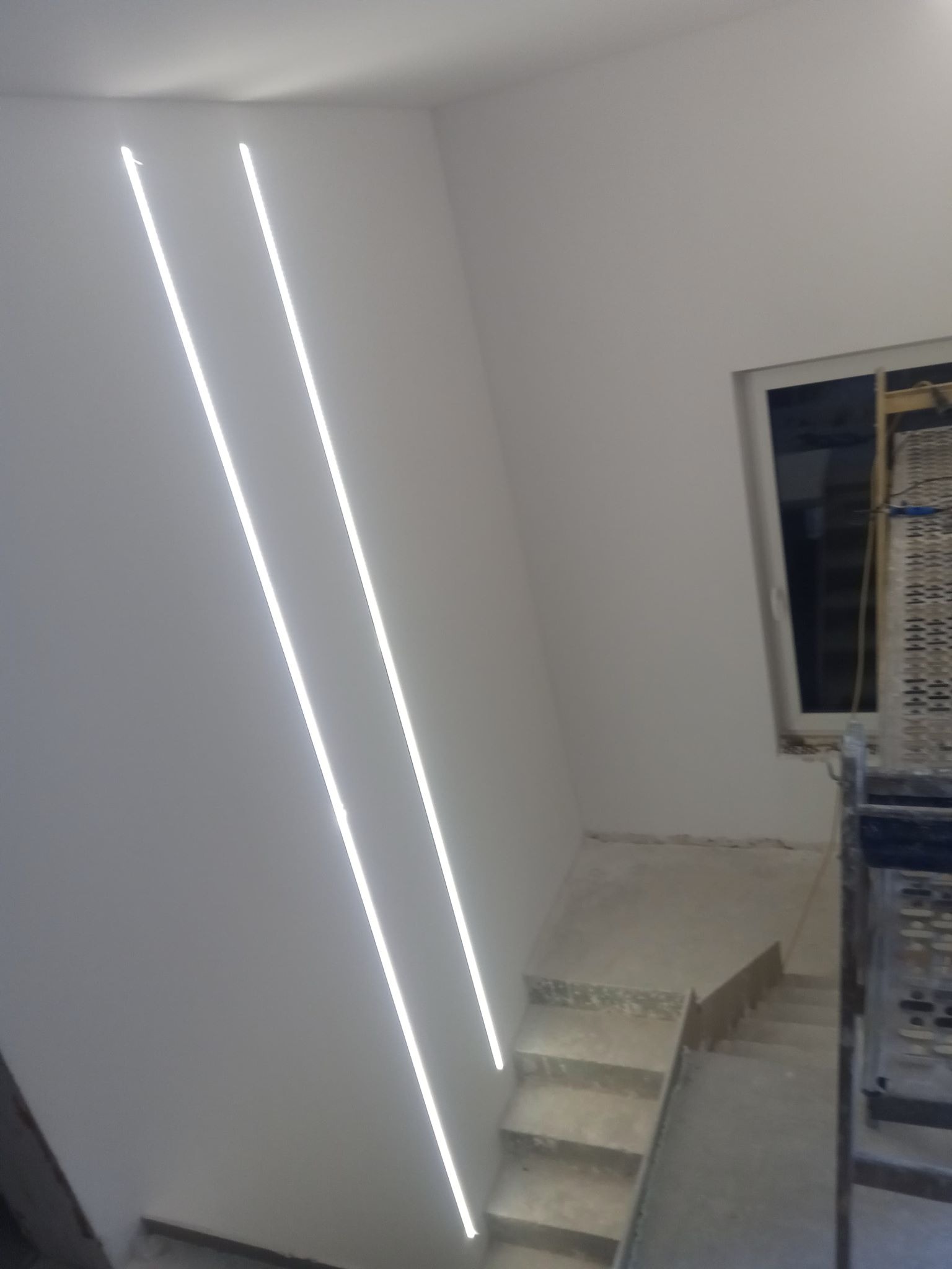 Klatka schodowa w trakcie wykończenia z pionowymi, liniowymi lampami LED wpuszczonymi w ścianę, widoczny fragment rusztowania przy oknie.