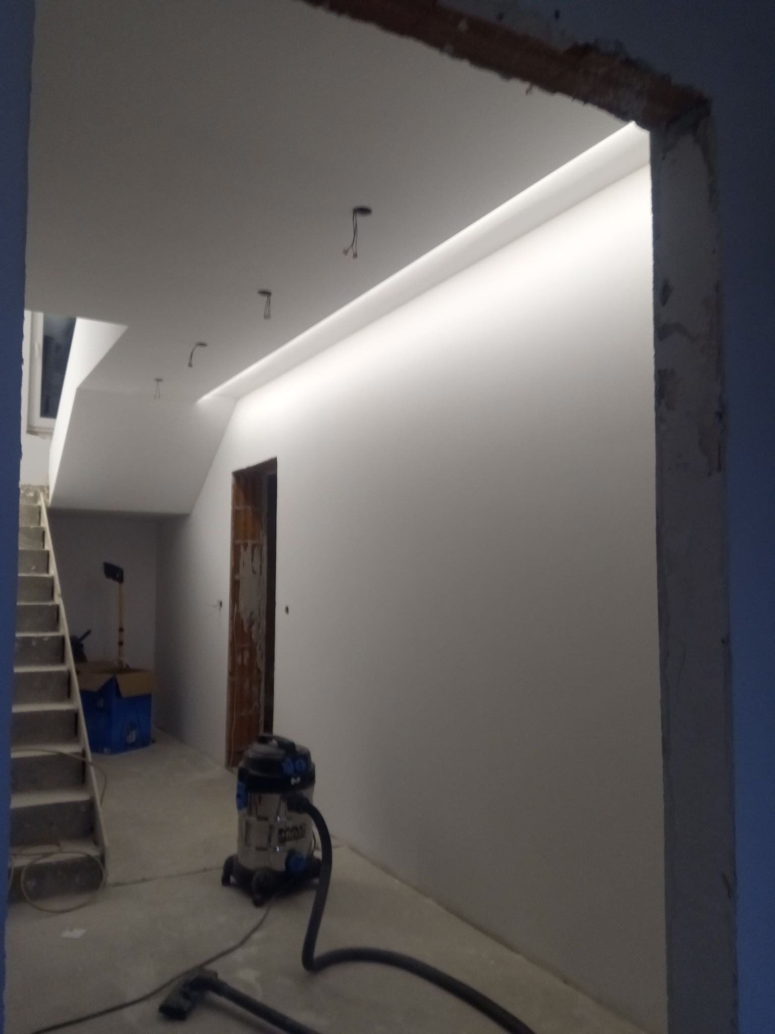 Wnętrze domu w trakcie remontu, widoczne schody, ściany po gładzi, wnęka z oświetleniem LED, otwarta futryna drzwi oraz odkurzacz przemysłowy na podłodze.