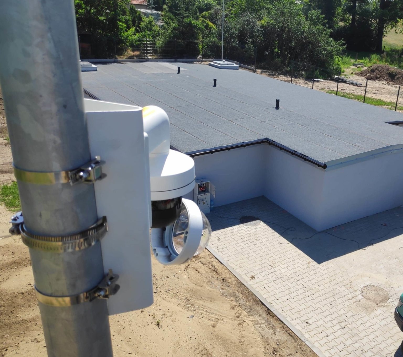 Zewnętrzna kamera monitoringu zamocowana na metalowym słupie za pomocą metalowych obejm, widok z góry na budynek z płaskim dachem i kostką brukową.