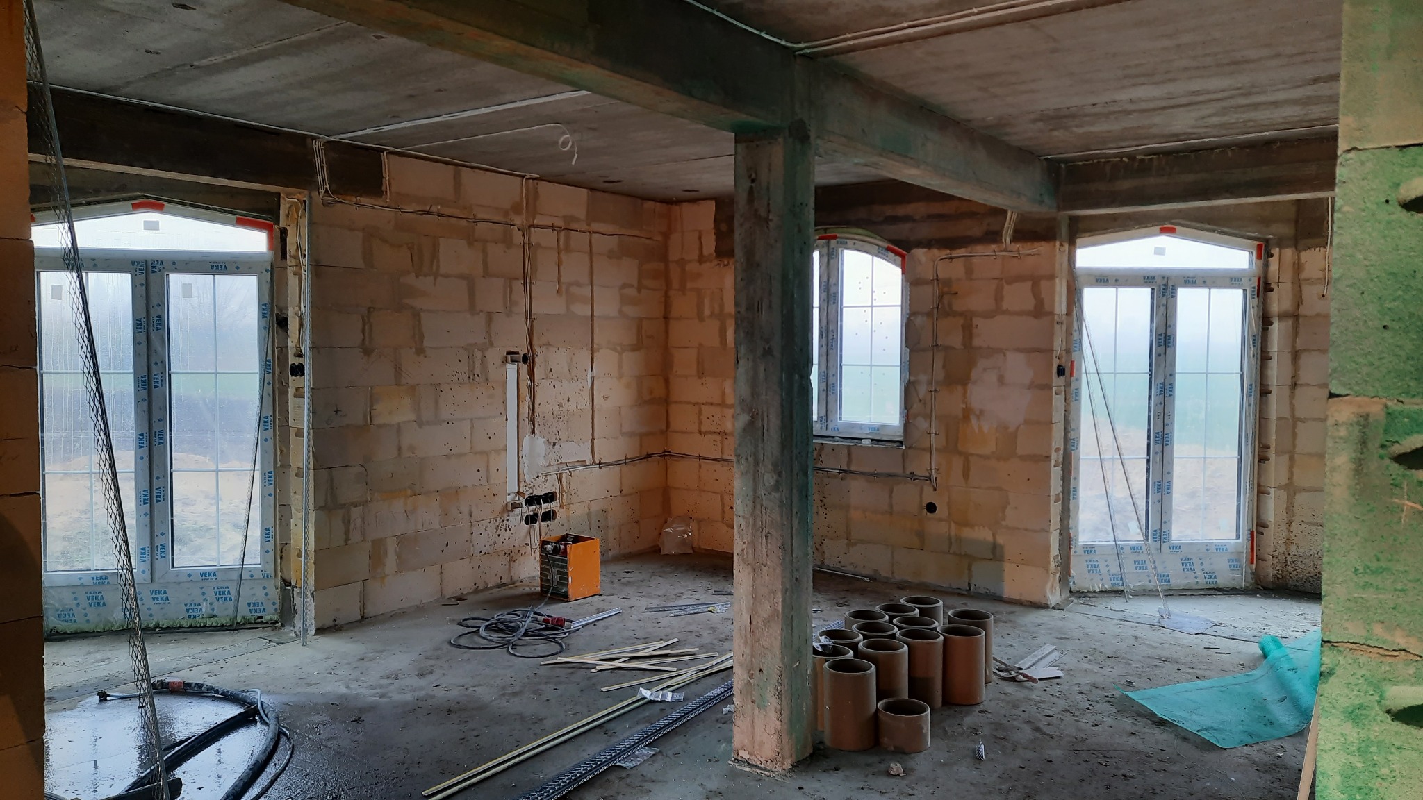 Surowy stan wnętrza z oknami PCV przed położeniem tynków maszynowych, widoczne przewody elektryczne i przygotowane rury do ogrzewania podłogowego.