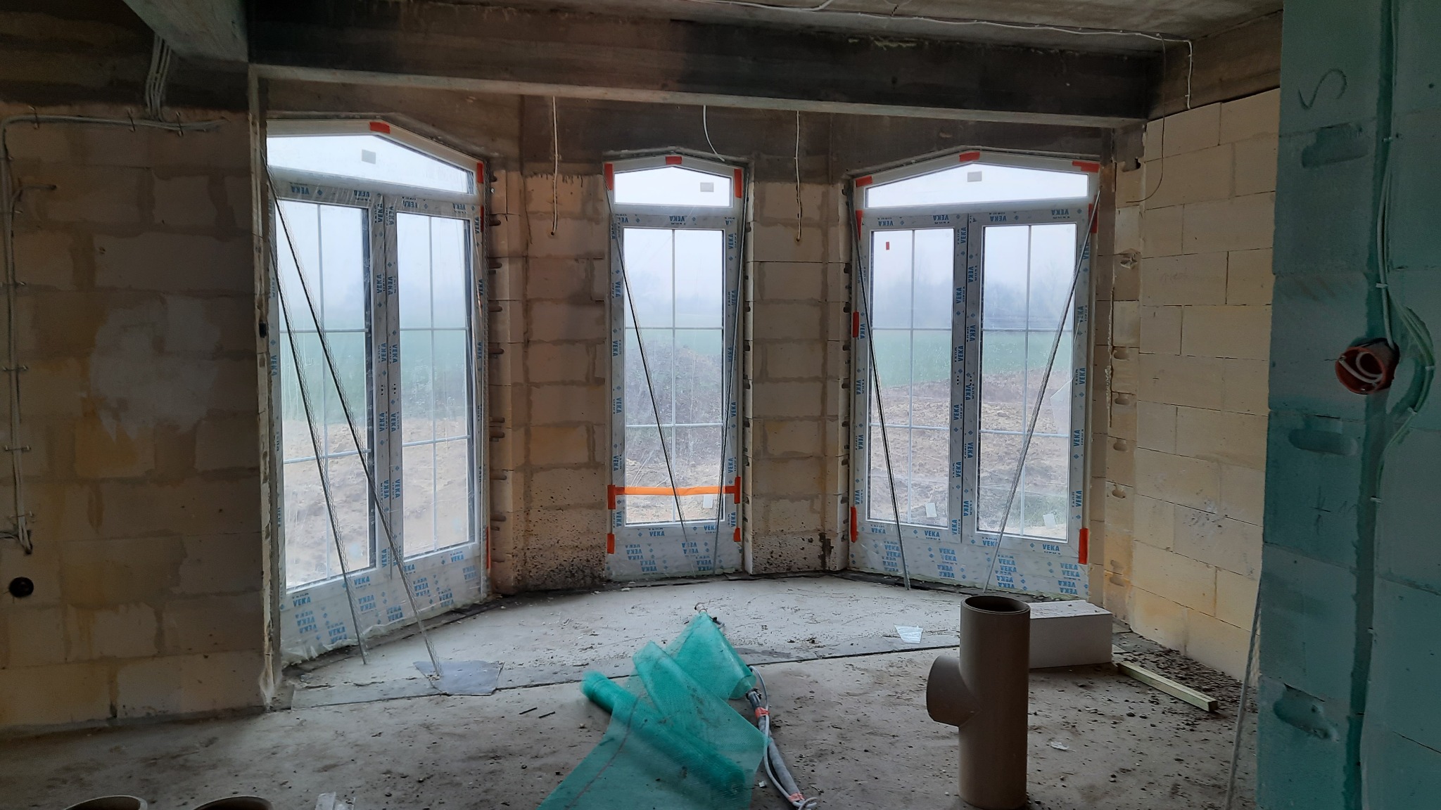 Surowy stan wnętrza z trzema oknami o nietypowym kształcie, widok na zewnątrz zasłonięty mgłą, ściany z pustaków, instalacja elektryczna w trakcie montażu, widoczne elementy instalacji...
