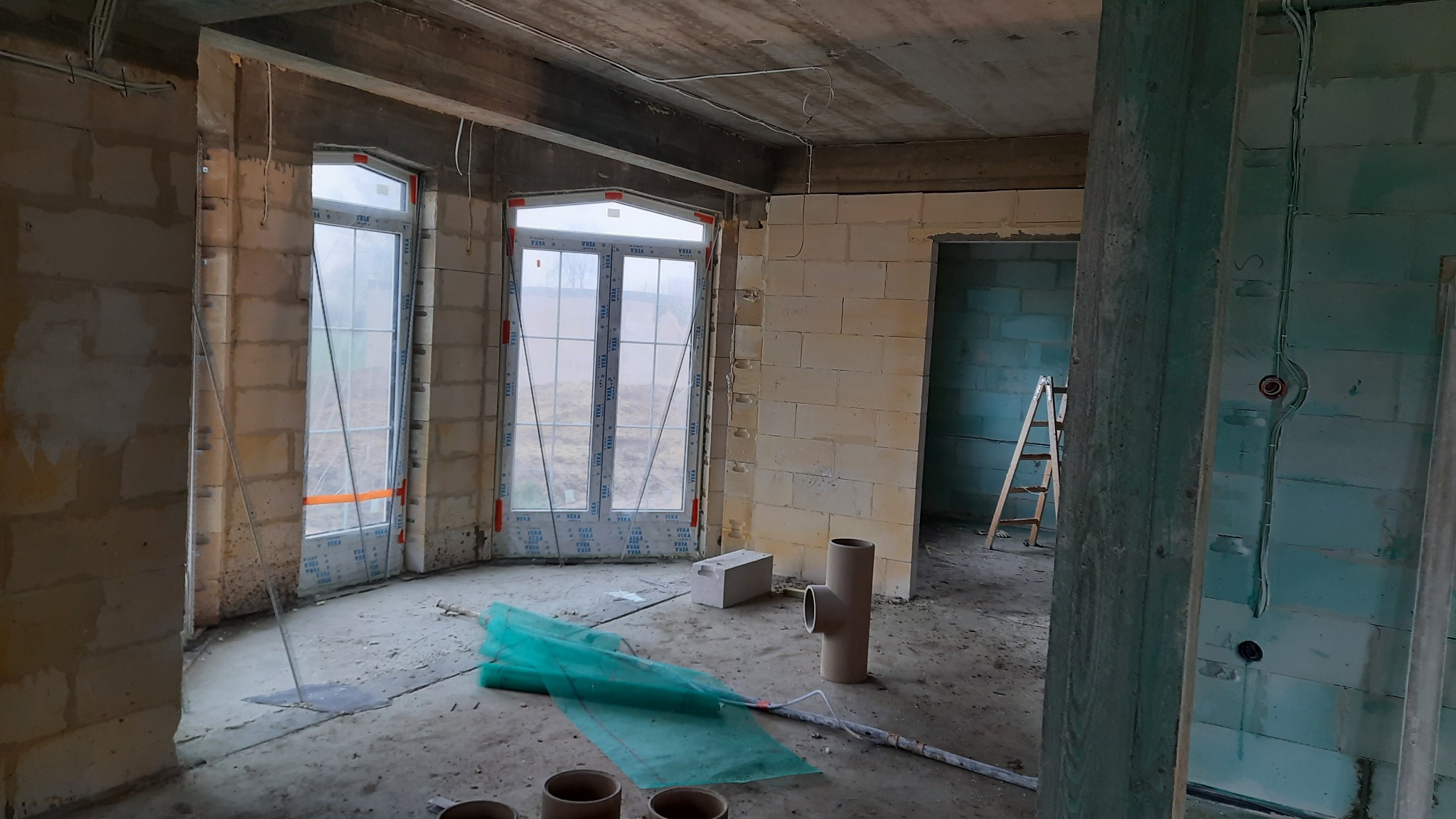 Surowy stan wnętrza domu z oknami Veka, widoczne bloki betonu komórkowego, instalacje elektryczne, rury kanalizacyjne i drabina w tle, przygotowane do dalszych prac wykończeniowych.