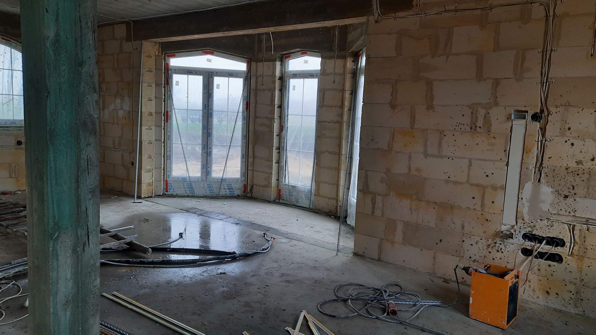 Wnętrze surowego budynku w trakcie prac wykończeniowych, widoczne okna z folią ochronną, ściany z bloczków, kable elektryczne i narzędzia, mokra posadzka.