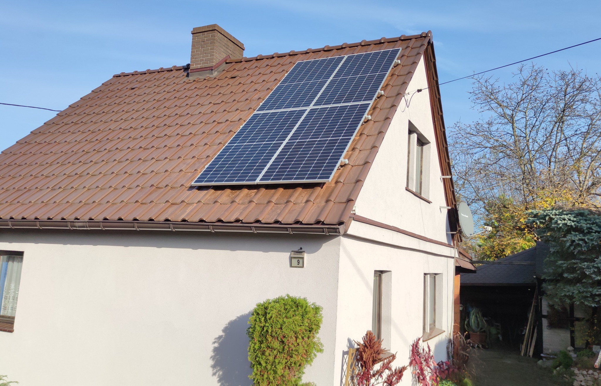 Instalacja fotowoltaiczna składająca się z czterech paneli na brązowym, spadzistym dachu białego domu jednorodzinnego w Rudzie Śląskiej, widok z boku na tle błękitnego nieba.