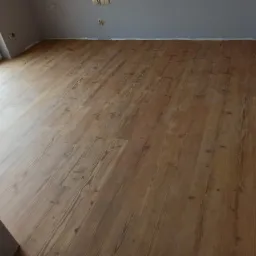 Ułożona podłoga z paneli o wzorze drewna sosnowego, widoczna w pustym pokoju z szarymi ścianami i białymi listwami przypodłogowymi, gniazdka elektryczne na ścianie.