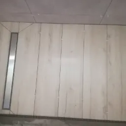 Ściana łazienki wyłożona jasnymi płytkami imitującymi drewno z zamontowanym odpływem liniowym, widoczne krzyżyki dystansowe w fugach.