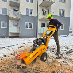 Traw-mat Usługi ogrodnicze Mateusz Szkaradek - Pracownik w kasku obsługuje frezarkę do pni drzew na tle budynku. Wokół leżą wióry i resztki po wycince. Żółta maszyna Liwex F-360 SW w akcji.