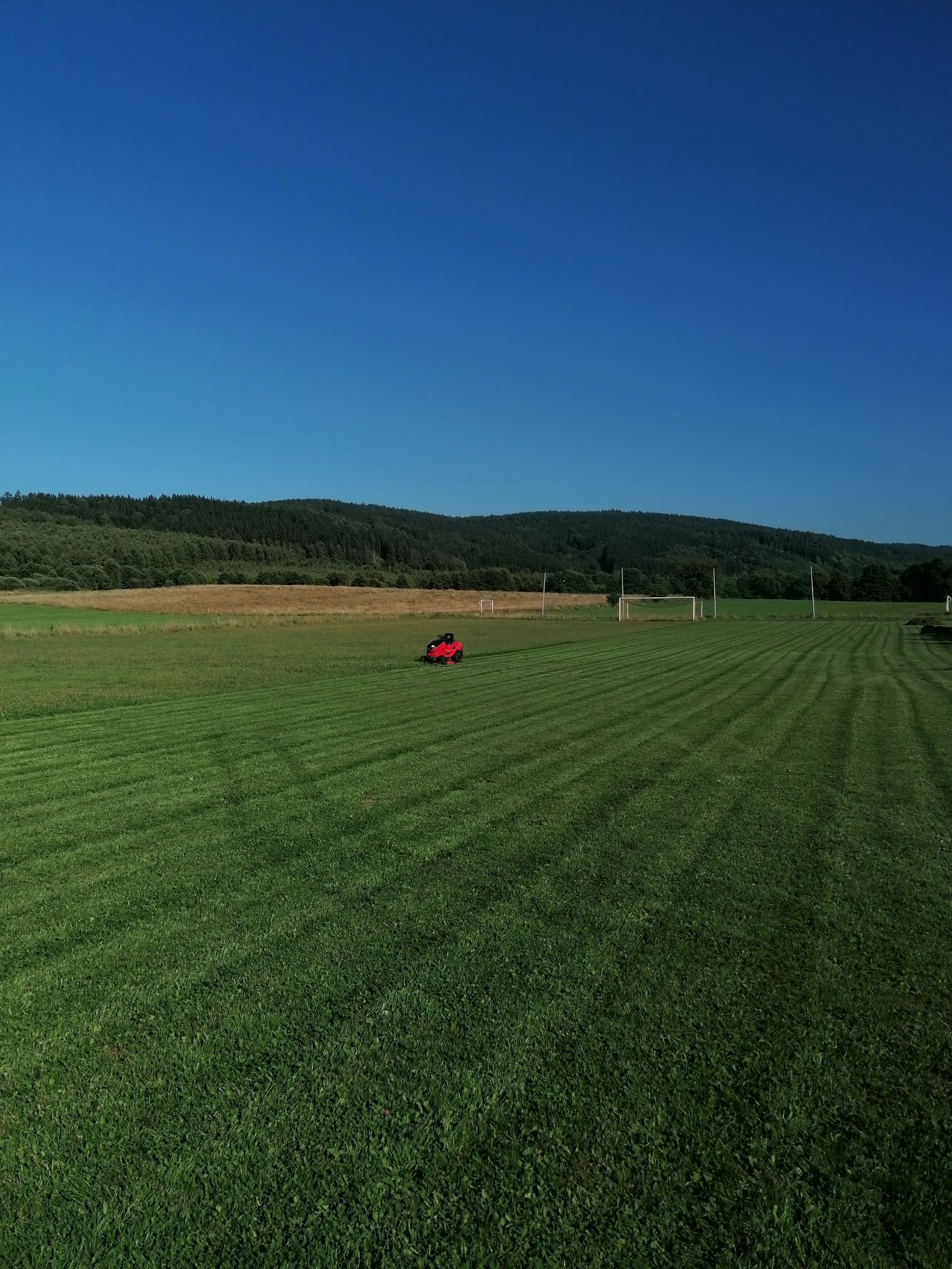 Czerwona kosiarka automatyczna w trakcie pracy na rozległym, zielonym boisku z bramkami, zalesione wzgórza w tle pod błękitnym niebem.