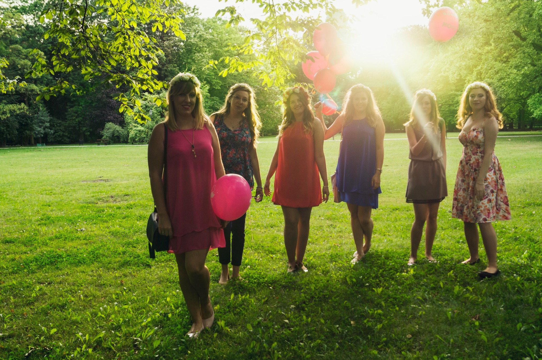 Grupa młodych kobiet w letnich sukienkach i wiankach na głowach pozuje na zielonej łące, trzymając różowe balony; słońce prześwieca przez drzewa w tle.
