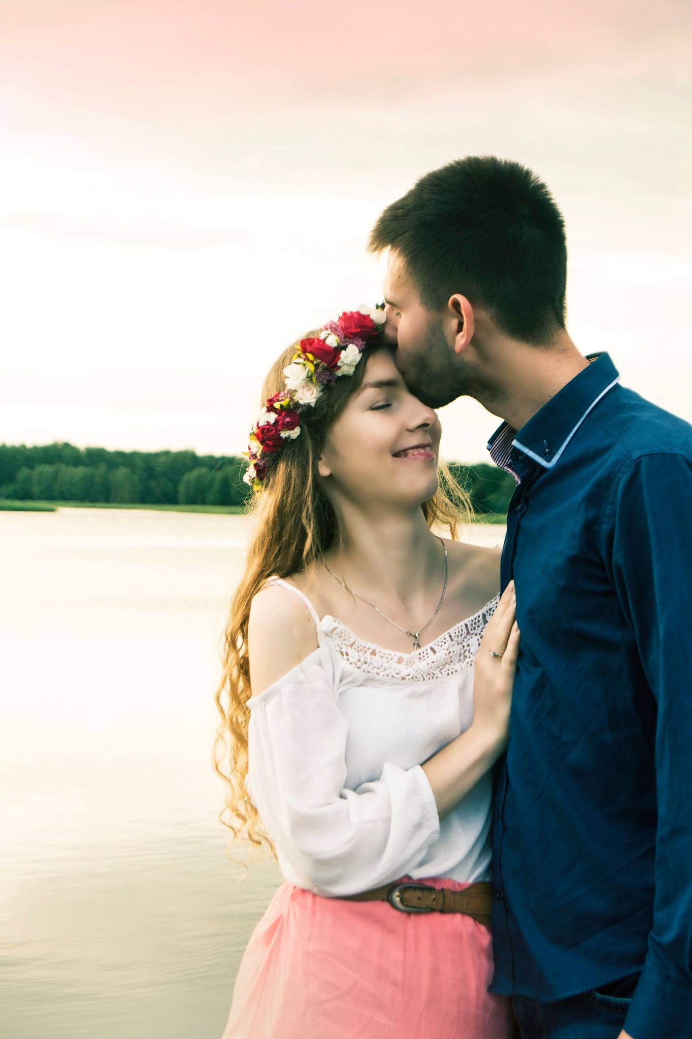 Mężczyzna całuje kobietę w wianku na tle jeziora; kobieta w białej bluzce z koronką i różowej spódnicy, on w granatowej koszuli.