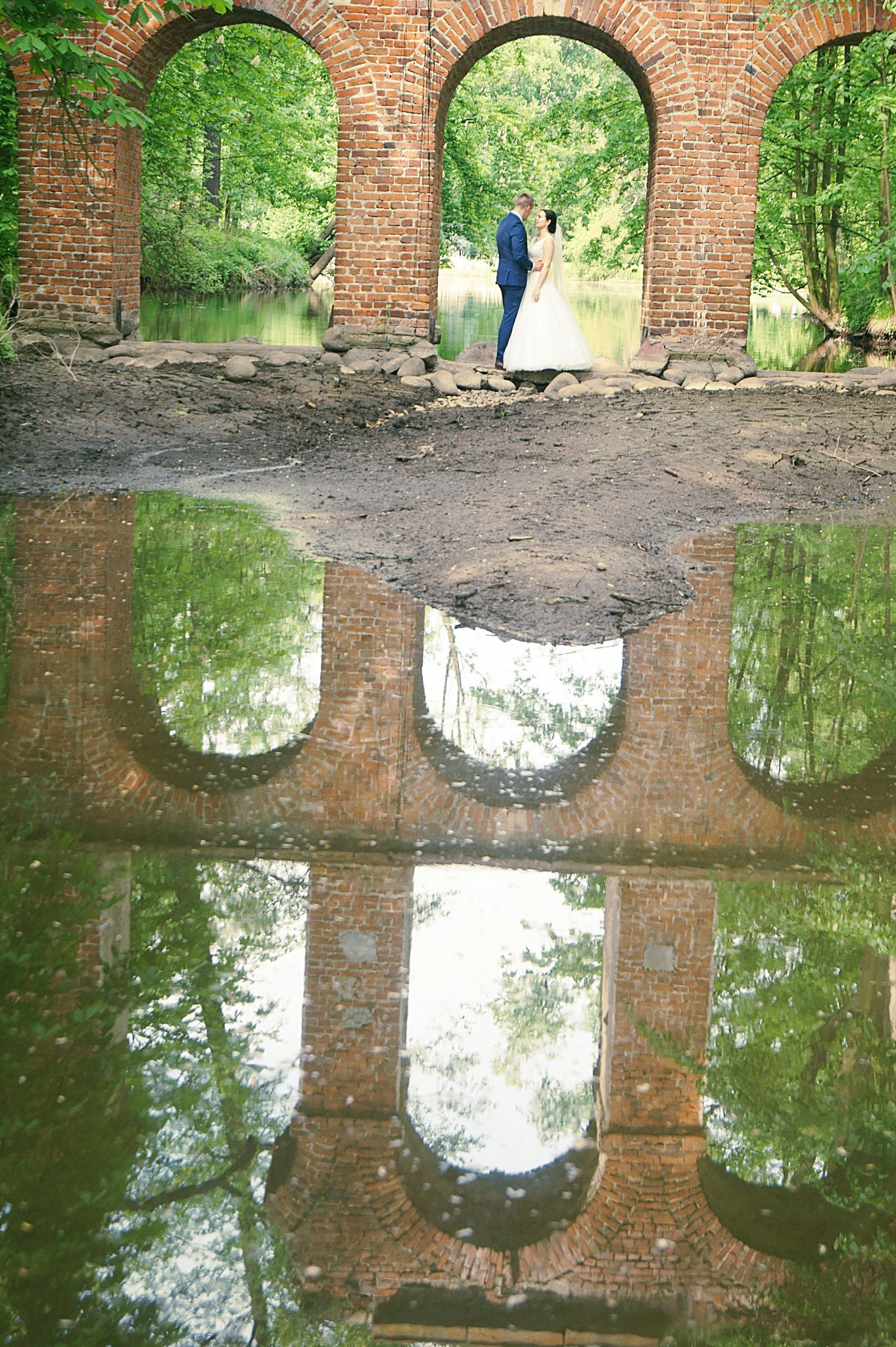 Para młoda stoi w prześwicie ceglanego mostu, którego odbicie w kałuży tworzy symetryczną kompozycję z zielenią drzew w tle.
