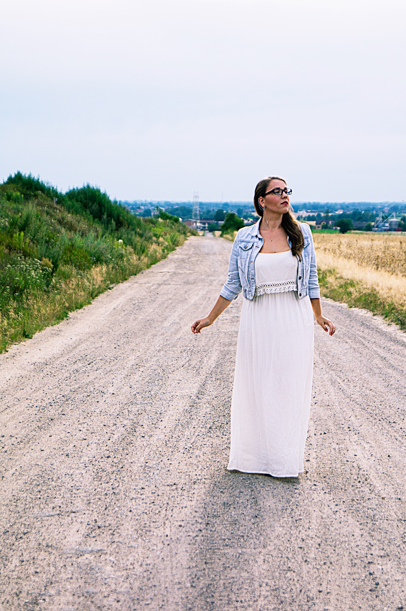 Kobieta w białej sukience i jeansowej kurtce spaceruje po polnej drodze, wzrok skierowany w dal, w tle widoczne zabudowania i słupy wysokiego napięcia.