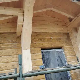 Drewniana konstrukcja domu w Glinczu, częściowo zabezpieczona folią, z widocznym rusztowaniem i narzędziem elektrycznym na nim. Fragment ściany po prawej stronie z ciemniejszym odcieniem drewna.