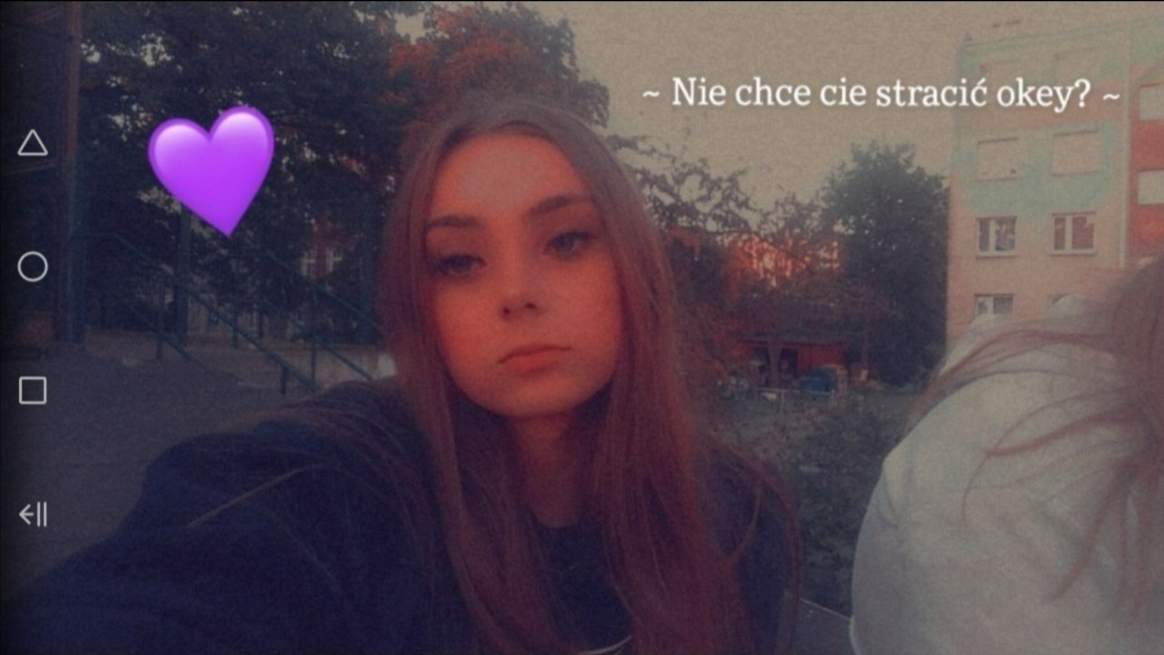 Selfie młodej kobiety z emotikonem serca, w tle blok mieszkalny i zieleń, na zdjęciu widoczne elementy interfejsu telefonu.